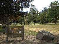 In the forefront is a large rock along with a sign displaying the name, management, and hours of operation for the park; in the background is a lawn and scattered trees.