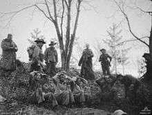 Five Asian soldiers sitting on a hillside with their hands behind their heads, while above them stands a group of Caucasian soldiers wearing slouch hats