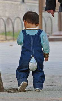 A walking boy, seen from behind, with a diaper visible through the open seam in the buttocks area of his denim overalls