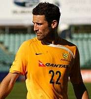 A young, dark-haired, bearded man in a yellow, green and white football shirt looks to his right, the sun casting a shadow across his body.