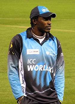 A man standing with his hands in his pockets. He is wearing a blue–black hat and top