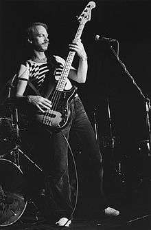 Brown is shown in a full length, black-and-white shot. He is leaning backwards and towards his right, with the bass guitar held at a steep angle. A microphone is in shot in front of him.