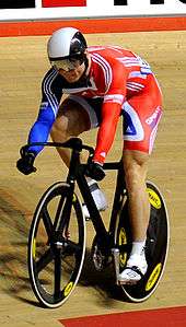 Chris Hoy wearing a bicycle helmet, visor, cycling shorts and top cycling on a racing bike in a velodrome.