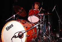 Chris Layton playing a drum kit. He has dark hair and is looking upwards.