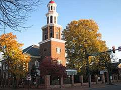 An 18th-century red brick church with white steeple behind a modern road in autumn.