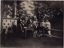 A black-and-white, slightly blurred photograph of a party outside a white house. To the left is a terrace, to the right a bench and in the middle a small table. There are many persons in the picture, who are formally dressed.