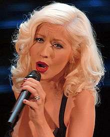 A picture of a blonde haired woman, singing while looking straight to the camera