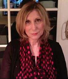 A low-quality photo of Christina Hoff Sommers, sitting in what appears to be her home, wearing a dark red shirt and red and black scarf.