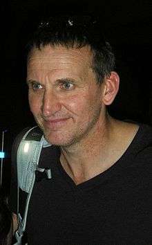 An upper-body shot of a middle-aged Eccleston with short dark hair, wearing a dark top, with the white strap of a bag slung over his right shoulder.