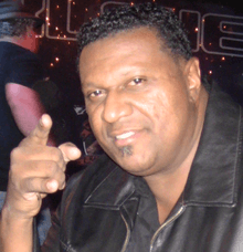 A man with curly hair, wearing a black leather jacket and pointing his finger. A man wearing a black shirt is standing in the background.