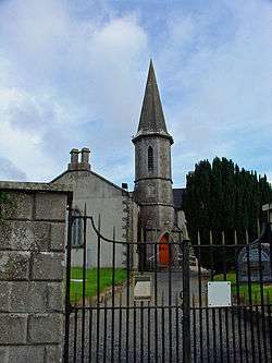 Behind a wrought iron gate sits a church with grey stone quoins. An octagonal steeple with a tall pointed roof adjoins the right side of the nave and contains a ground-level entry with red door.