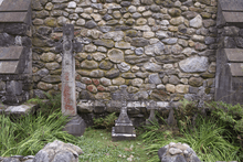 Several gravestones between buttresses against a stone wall. The one in the right is taller, narrow with some ornate decoration and a red area near the bottom. The others are small crosses