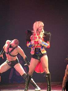 Image of a blond woman. She wears a red circus ringleader jacket with feathers, shorts and stockings. She holds a whip around her neck. In the woman's left, there's a man dressed in S&M bondage clothing.