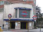 A light-grey-tiled building with a blue sign reading "CLAPHAM SOUTH STATION" in white letters and a woman walking in front wearing a black jacket