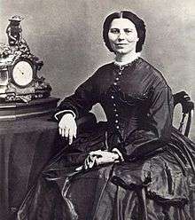 19th century photograph of a woman seated with her arm resting on a table. Her dark hair is neatly partedin the middle. She is smiling slightly