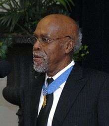 Clarence Sasser, a gray-haired black man standing at a microphone, wearing a medal on a blue ribbon around his neck.