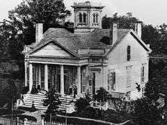 A white house with multiple chimneys and a tower.  People are on the front porch and someone is arriving at the home and about to embark up the staircase. The front porch has a roof over a set of four columns. Trees are visible on the lawn.