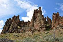 A row of rock spires connected at the base rises toward a partly cloudy blue sky.