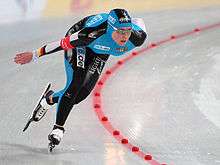 A female speed skater turns around a curve. She is wearing a black and blue body suit.