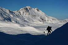 A hiker ascends a snowfield, with the sunlit slopes of a large, snowy mountain in the background
