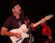 A man wearing a short-sleeved black shirt playing a white guitar and singing into a microphone. The background is black, except for a seated man playing on a keyboard and singing into a microphone.