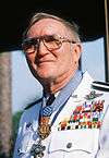 Head and shoulders of an older white man with glasses, wearing a white military jacket with a very large array of ribbon bars on the left breast and two medals hanging from ribbons around his neck.