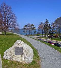 A gravel walkway curves past a whitish stone in which a historic marker has been embedded. In the background you can see a road and then water beyond.