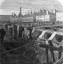 drawing showing a large quantity of collapsed brickwork retaining wall and destroyed timber scaffolding alongside a flooded excavation. Beyond the cutting is a wide open area with small figures inspecting the damage. The backdrop is formed by a collection of two-storey buildings many of which have large chimneys.