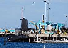 A submarine next to a dock, which is occupied by several cranes and other mechanical equipment