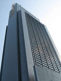 Ground-level view of a building with a rectangular cross section. Two sides of the tower are visible; one has a concrete, windowless facade while the other's facade is a glass curtain wall.