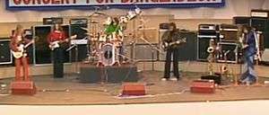 Five men are on stage with the man at left playing a guitar and facing to his left. The second man plays a bass guitar. The third man is behind his drum kit. The fourth man is playing a guitar and the last man is playing saxophone.
