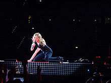 A female blond performer sitting on all fours on a stage. She is singing to a microphone while holding it with her right hand. The background is dark but a red glow falls on the woman.