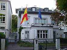 A two storey white building with an attic. Two flagpoles, one with the flag of Spain, the other with the European flag, are in front of the building.