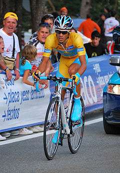 A cyclist in a gold jersey with blue trim and blue shoes riding down a road, with spectators watching him from behind a guardrail and one headlight from a car behind him just in the frame