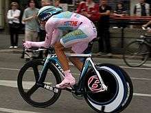 A cyclist wearing a pink skinsuit and shoes, sitting crouched in an aerodynamic position over his bicycle. Spectators watch from the roadside.