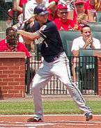 right-handed baseball player awaits a pitch at home plate with his left leg cocked to stride for a swing. The bat is over his right shoulder and he is wearing a blue jersey and dark batting helmet.