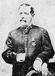 A white man with a mustache standing with his left arm resting on an object to his side and his right hand inside his jacket. A star-shaped medal is hanging from a ribbon on his left breast.