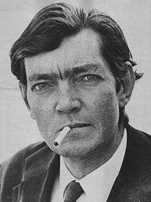Head shot of a dark haired man in a suit and tie with a cigarette in his mouth. He is looking at the camera quizzically.