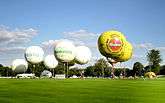 Several white balloons and two yellow balloons taking off from a grassy field.