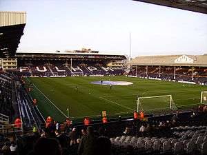 A colour photograph of Craven Cottage football stadium, showing the pitch in the centre of the image, surrounded stands. At the top of the photo, the word 'Fulham' is spelt out in white lettering on a the black seats.