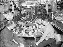 Around 15 men are drinking from white mugs at a table in a narrow compartment. They are surrounded by bunk-beds and machinery. The space is cramped and the men are packed closely together.