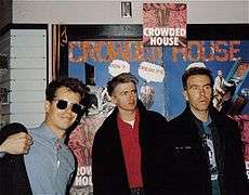 Three men are standing in front of posters advertising the band. Man at left is wearing sunglasses, smiling and adjusting his dark jacket. Man in middle is staring to his left and wears a similar dark jacket. Third man is also staring to his left and has a dark jacket.