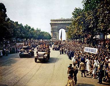 Crowds line the Champs Elysees as tanks and half tracks roll down it. The Arc de Triomphe is in the background. People are holding a sign that reads: "Viva de Gaulle".