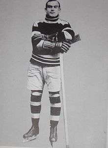a black and white photo of a young hockey player standing on his skates with his arms folded leaning slightly on an upside down hockey stick