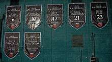 Seven large black shield-shaped banners are hung on a green wall, with white text for the name and number, or role that the individual played.