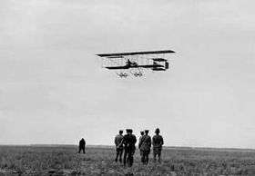 Biplane flying low over field, watched by a group of men