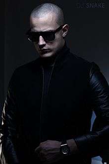 A man with a shaved head wearing black sunglasses, dark clothing and a watch