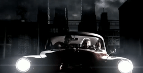 A blonde woman driving a vintage car at night in a city-like setting. She is accompanied by a male passenger who is gagged and tied-up.