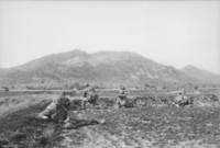 Soldiers wearing flak jackets and helmets are prone on an open field at the base of a large vegetated hill.