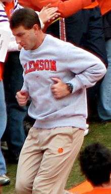 Coach Swinney in a grey sweatshirt and khaki pants, while running, with persons at rear.